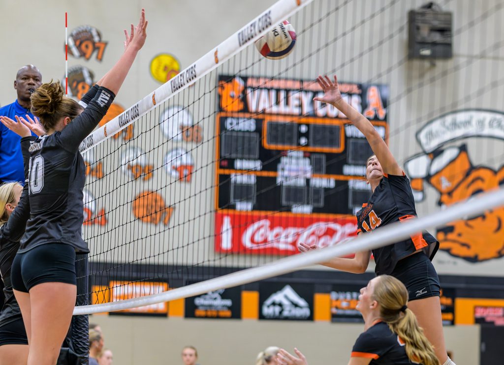 Inside a high school gymnasium, a volleyball player jumps to spike the ball over the net while opponents prepare to block.