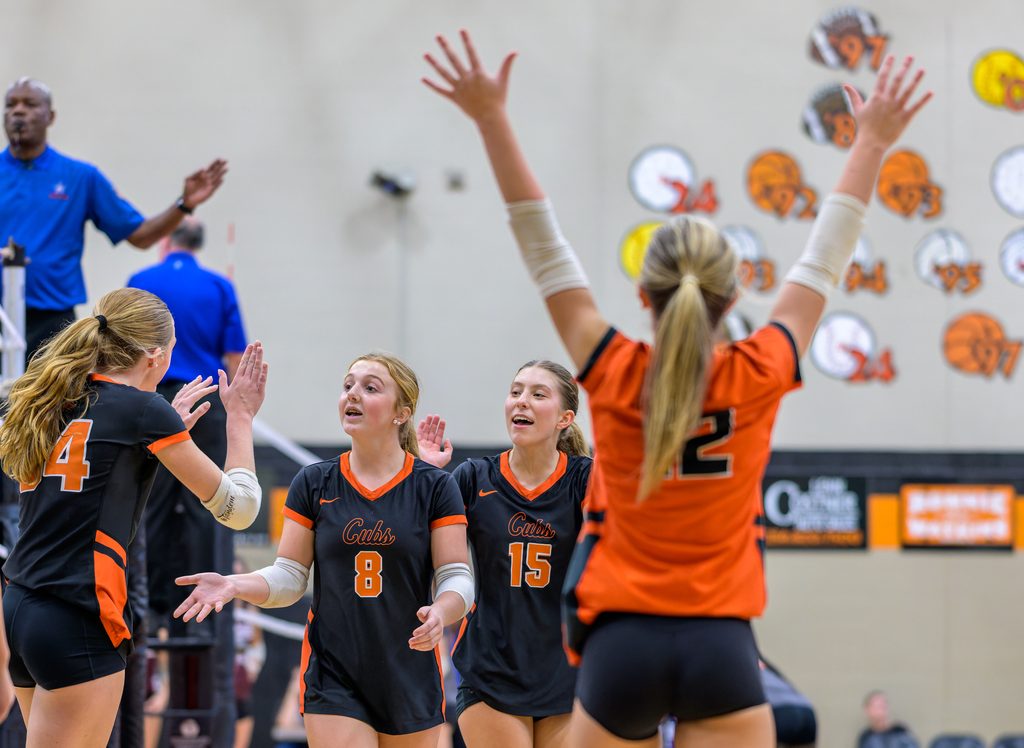 In a brightly lit high school gymnasium, several volleyball players wearing matching team uniforms celebrate joyfully on the court.