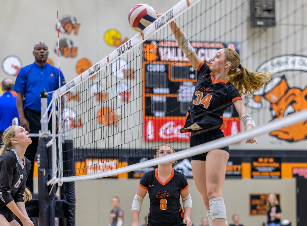 Inside a high school gymnasium, a volleyball player leaps to spike the ball over the net while teammates and an official watch.