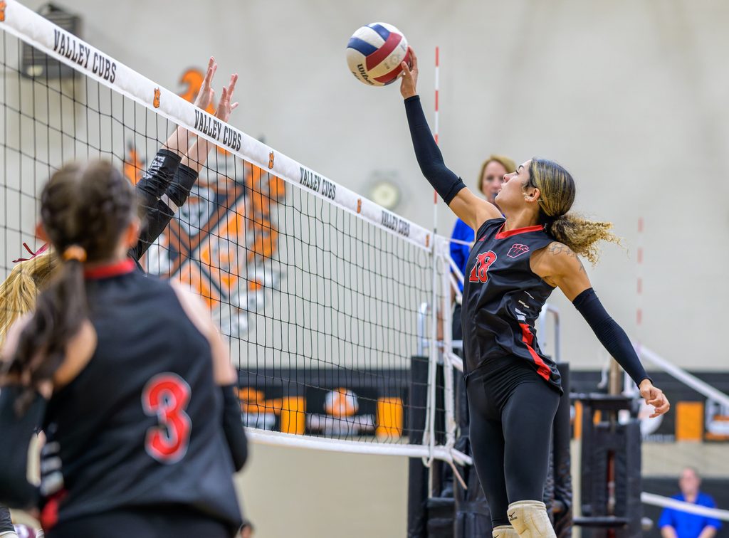 In an indoor high school gymnasium, a female volleyball player reaches high into the air to spike the ball over the net as opponents attempt to block.