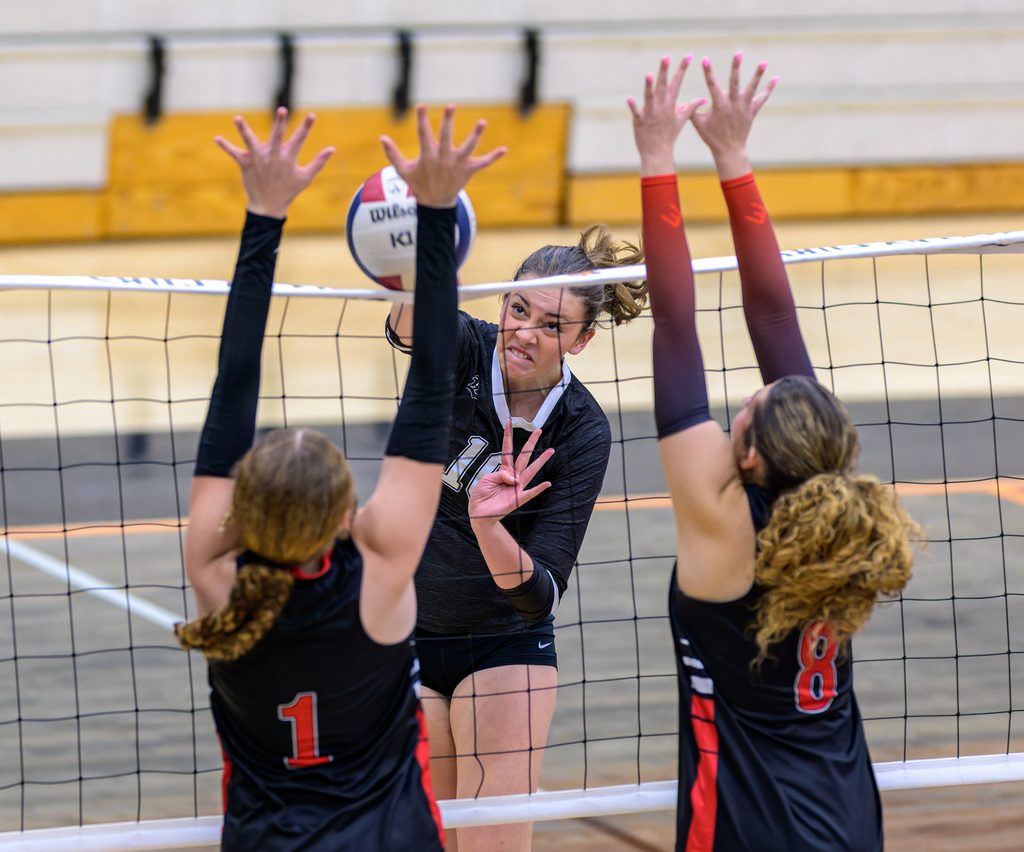 Inside a high school gymnasium, a volleyball player spikes the ball while two opponents jump at the net to block the play.