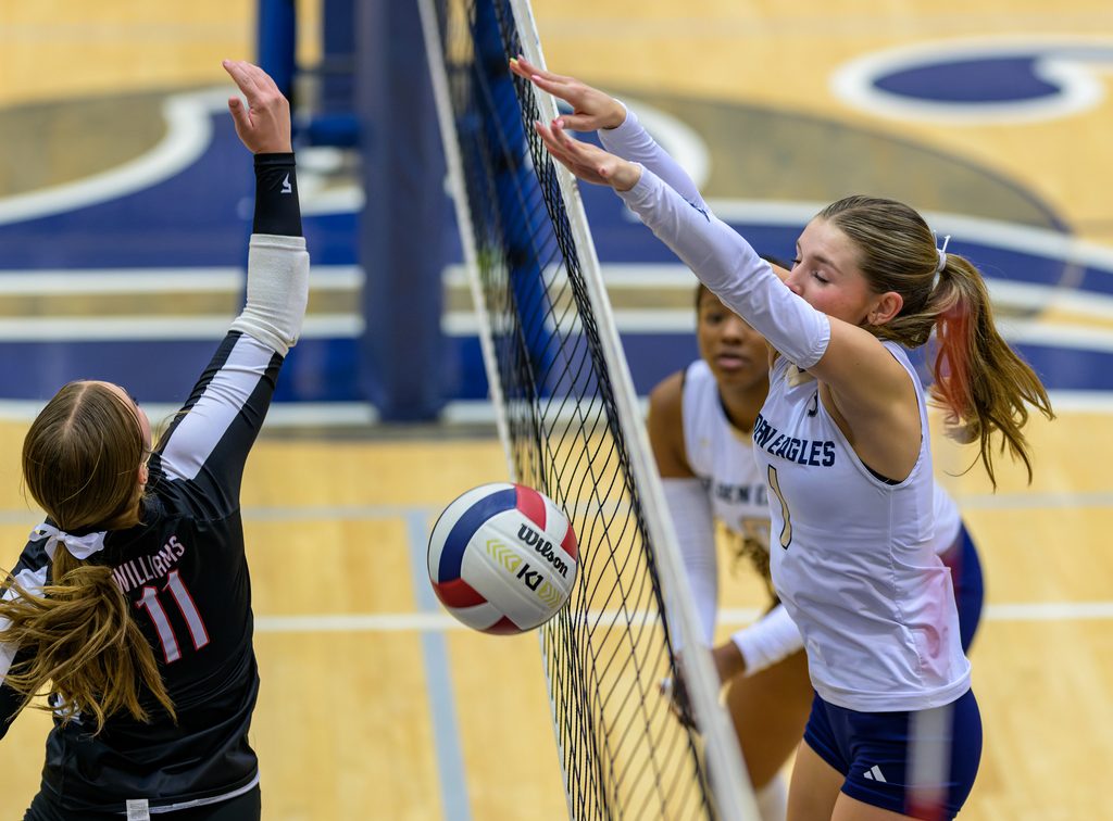Two high school volleyball players battle at the net during an indoor match on a polished wooden gymnasium court.