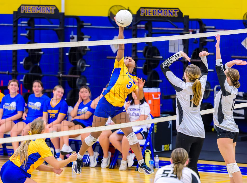 An indoor volleyball match is underway in a high school gym as a player in a yellow and blue uniform leaps to spike the ball over the net while opponents in white and black try to block.